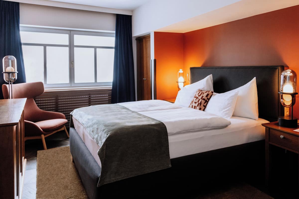 Una habitación de hotel con cama, escritorio, silla y ventana con cortinas.