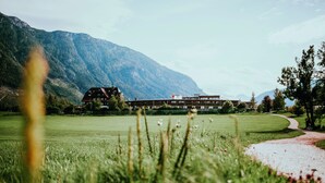 A building with a sloped roof, surrounded by a grassy field and mountains in the background. - Die Wasnerin G'sund & Natur Hotel (Bad Aussee)