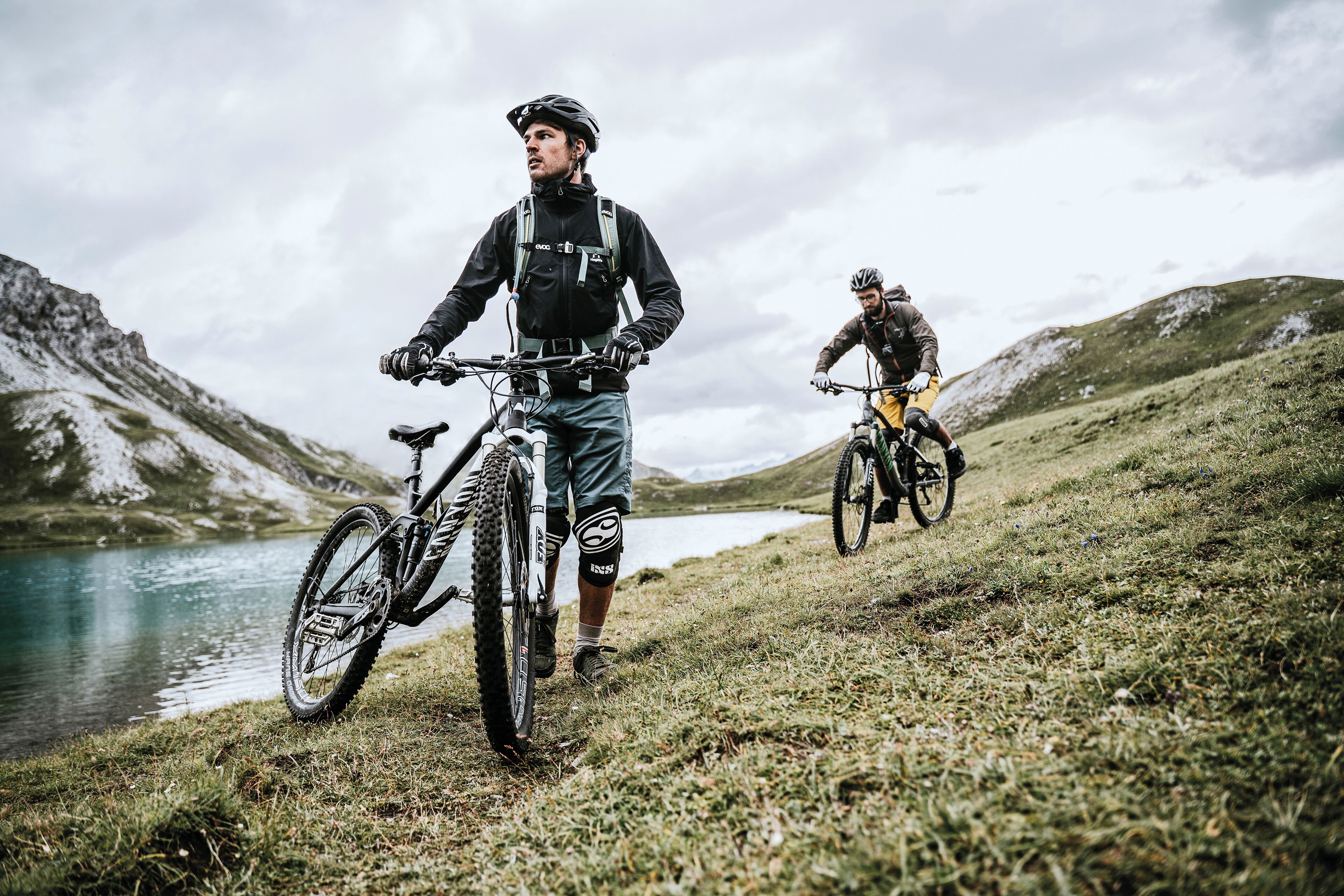 Zwei Radfahrer auf einem Bergpfad, im Hintergrund ein See.