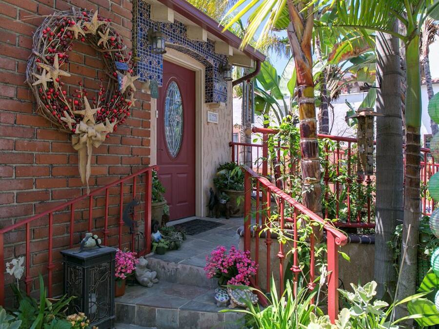 Een bakstenen huis met een rode deur, een rode trap en een tuin met diverse planten en een decoratieve krans.
