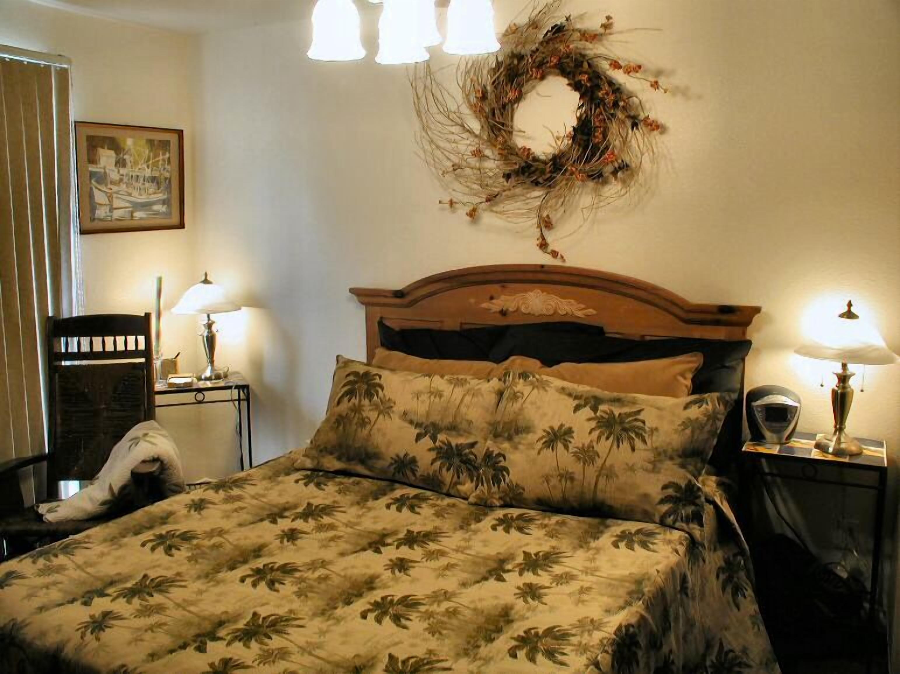 A hotel room with a bed, two bedside lamps, a chair, a framed picture on the wall, and a decorative wreath.