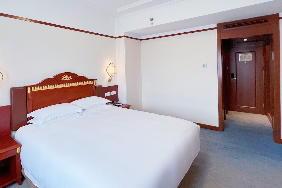 A hotel room with a large bed, a wooden headboard, a nightstand, a wall-mounted lamp, and a door leading to another room.