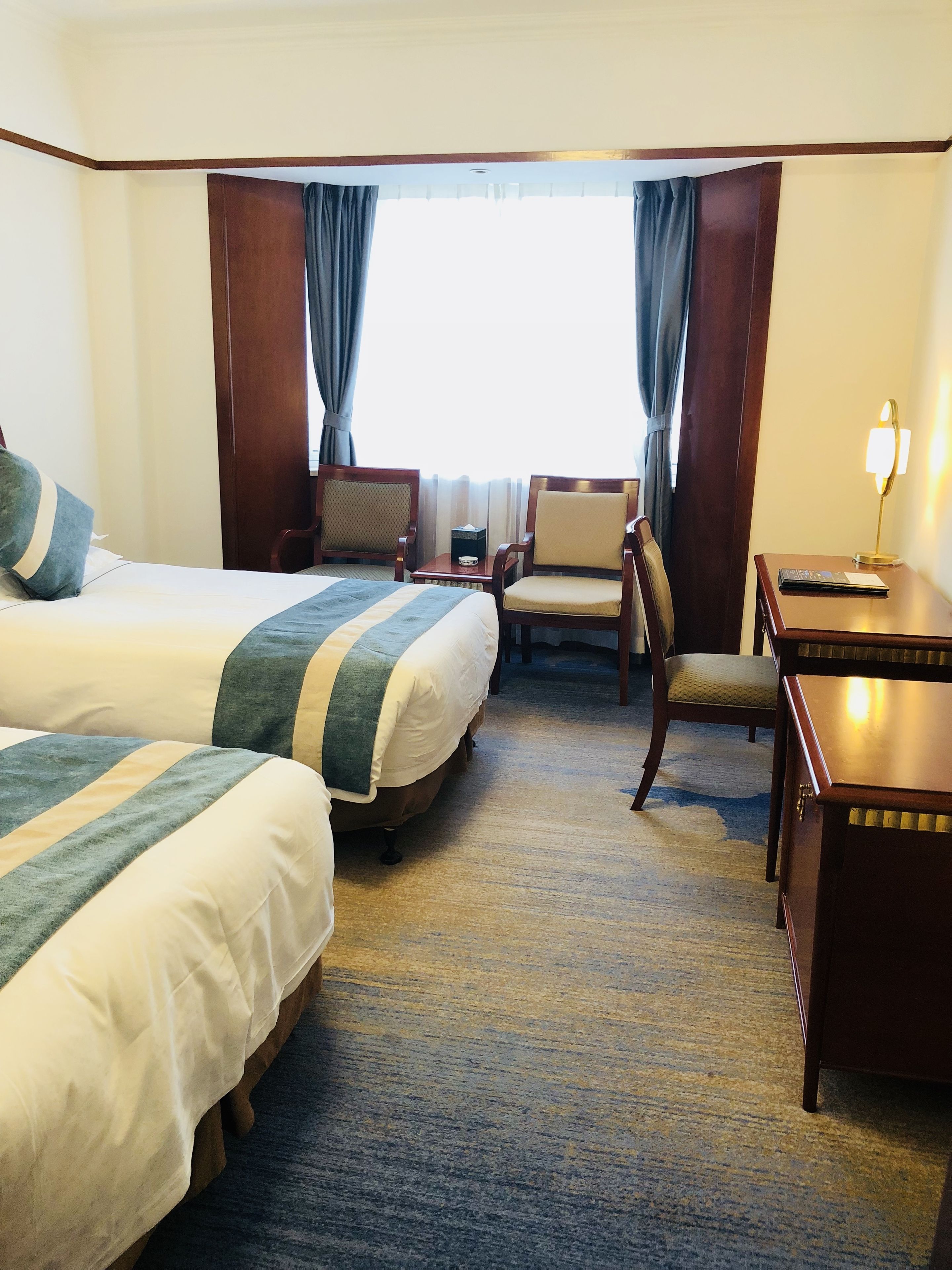 A hotel room with two beds, a desk, a chair, and a lamp.