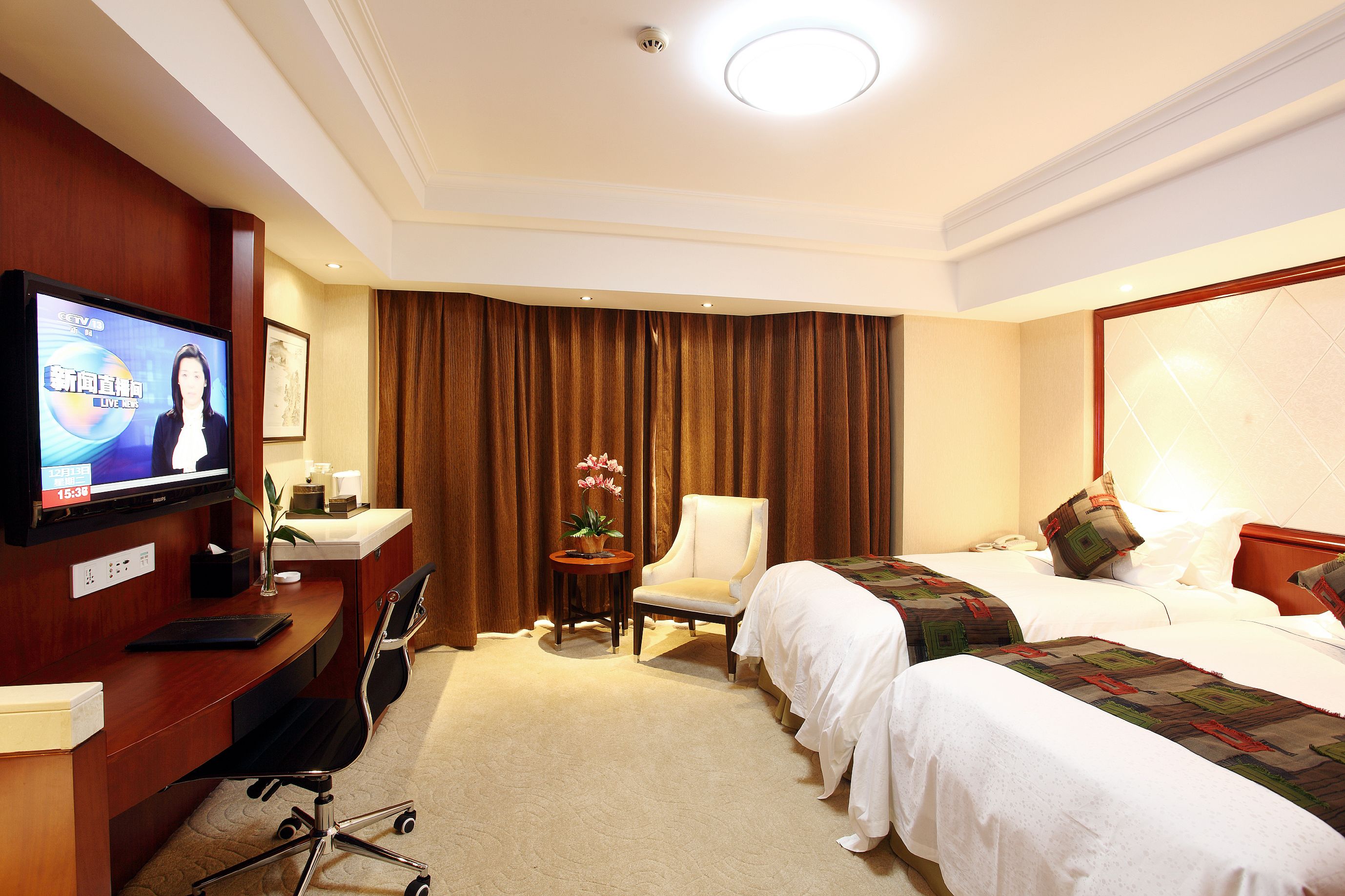 Ein Hotelzimmer mit Fernseher, Schreibtisch, Stuhl, zwei Betten und einem kleinen Tisch mit Pflanze.
