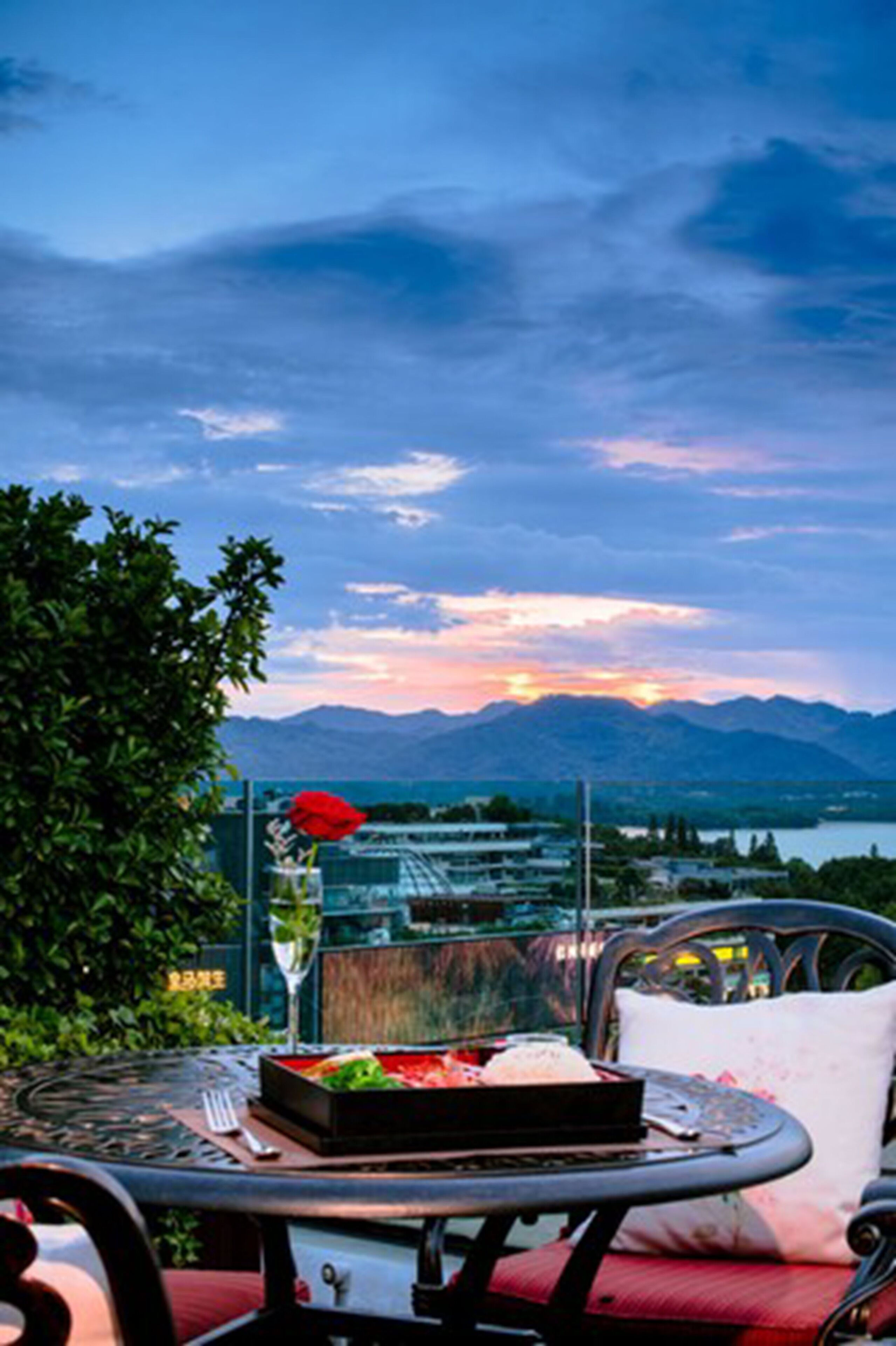 A dining table with a meal, a red umbrella, and a scenic view of mountains and a lake at sunset.
