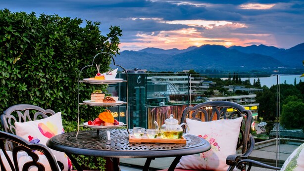 Un soporte de tres niveles para postres con varios dulces y una tetera sobre una mesa metálica redonda, con el paisaje urbano y las montañas al fondo durante el atardecer.