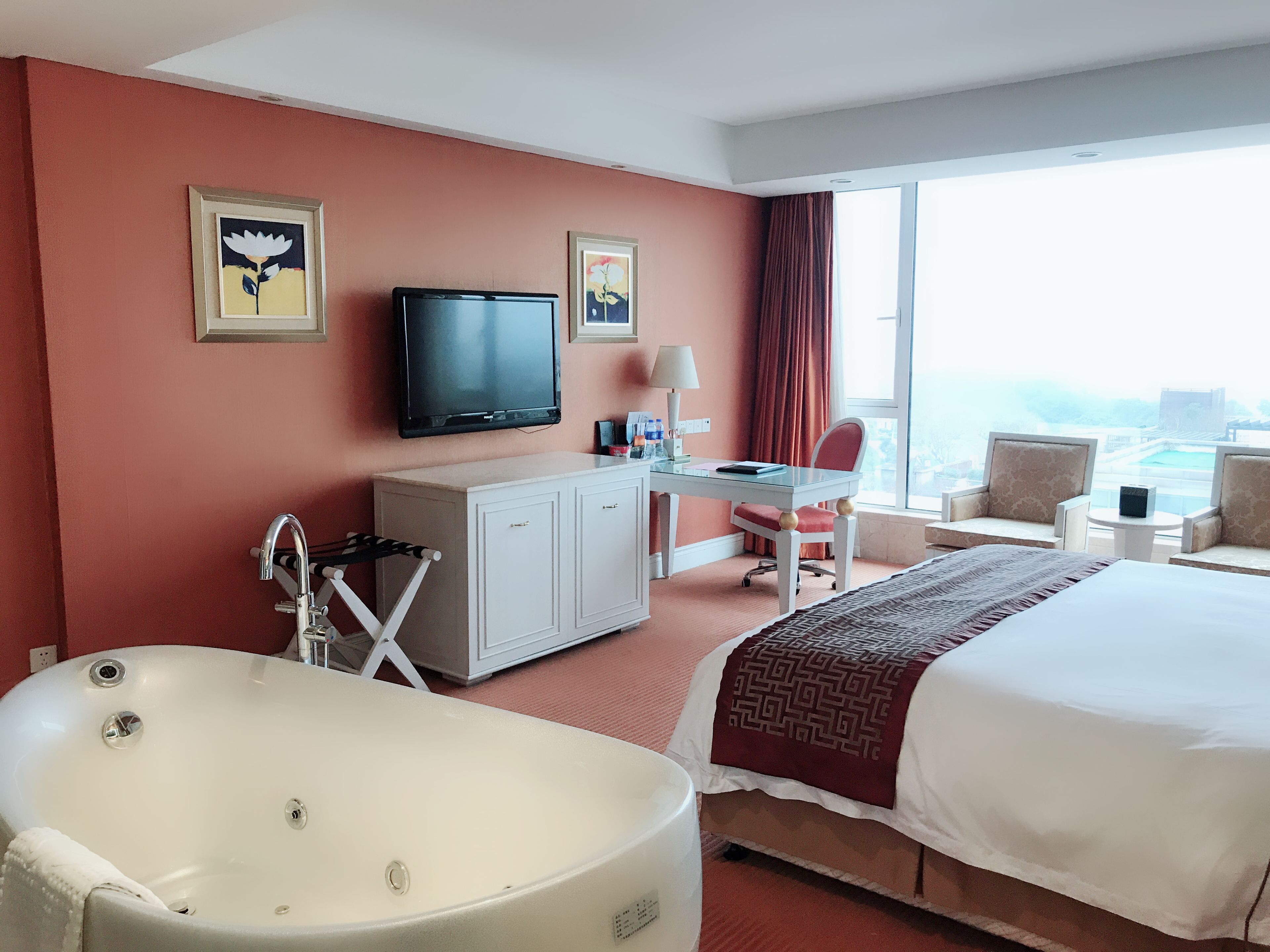 Habitación de hotel con una cama grande, un escritorio con silla, televisión, bañera y un ventanal con vistas a la ciudad.