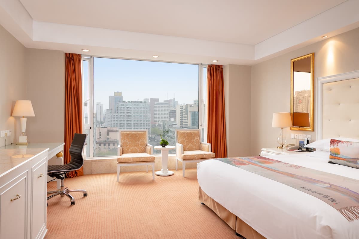 A hotel room with a large window, a bed, a desk, two chairs, a small table with a plant, and a mirror on the wall.