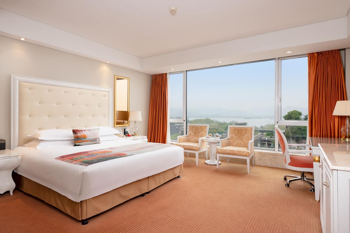 A hotel room with a large bed, two armchairs, a desk, and a view of the city through large windows.