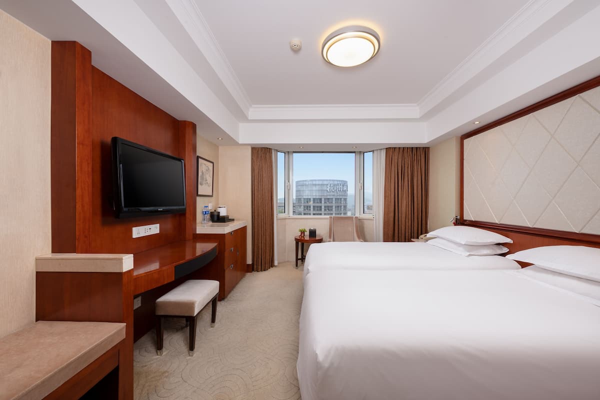 A hotel room with a large bed, a desk with a television, a small table, and a window with a city view.