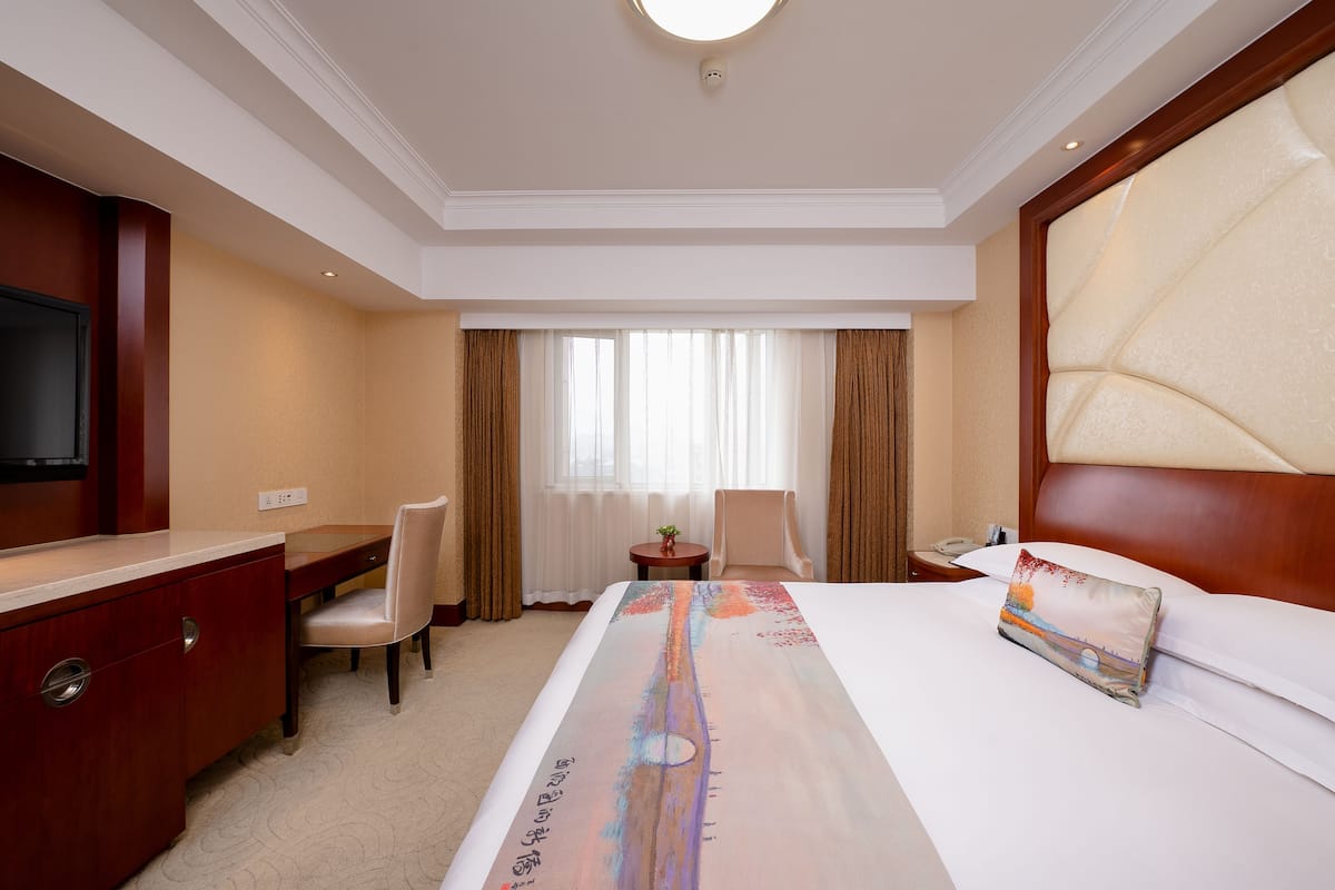 A hotel room with a bed, a desk, a chair, a small table with a lamp, and a TV.