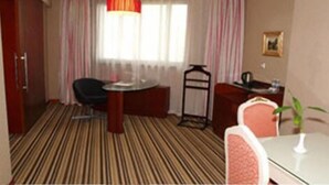 A hotel room with a striped carpet, a round table, a chair, a desk with a chair, a hanging lamp, and a window with curtains.