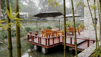 Una terraza de madera con sillas y mesas, rodeada de bambú y un muro de piedra.