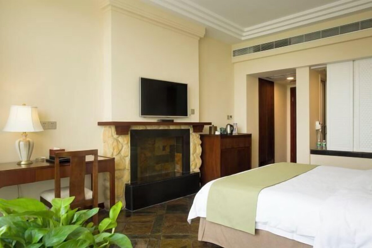 A hotel room with a bed, a television mounted above a fireplace, a desk with a lamp, and a plant.