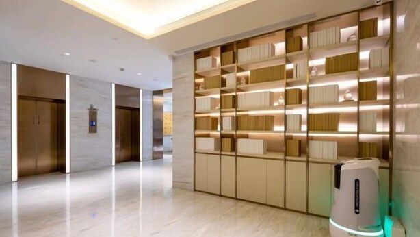 Un hall d’hôtel moderne avec un sol en marbre, un kiosque en libre-service et un mur recouvert d’étagères où les livres sont rangés avec soin.