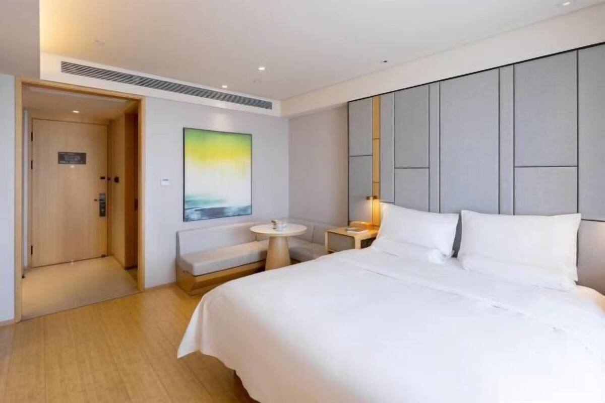 A modern hotel room with a large bed, a small round table, a bench, and a painting on the wall.