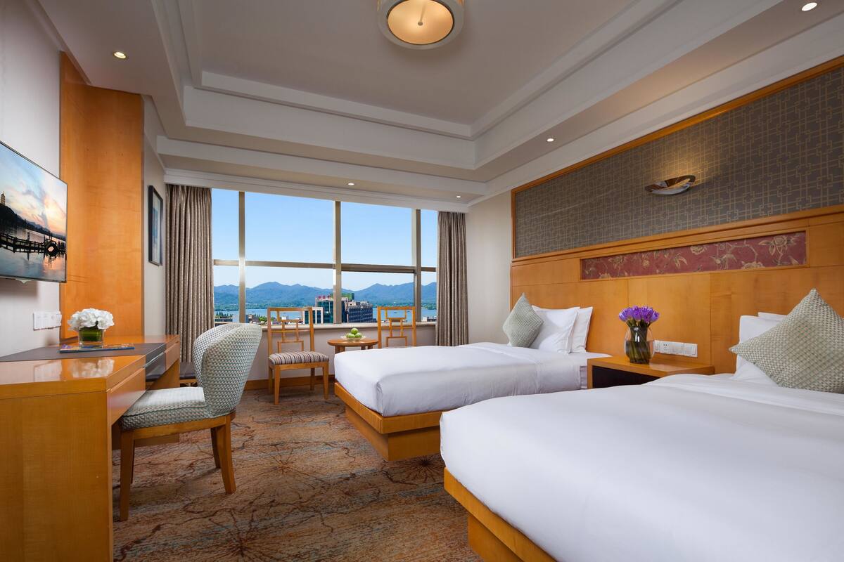 Ein Hotelzimmer mit zwei Betten, einem Schreibtisch, einem Sessel, einem kleinen Tisch und einem großen Fenster mit Bergblick.