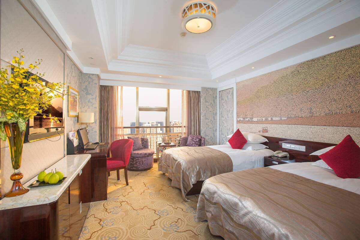 Ein Hotelzimmer mit zwei Betten, einem Schreibtisch, einem Stuhl, einer Vase mit Blumen und Blick auf die Stadt.