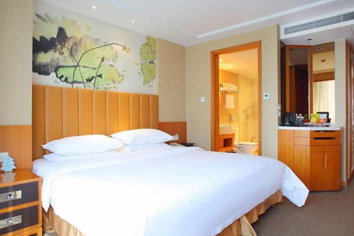 Une chambre d’hôtel avec un grand lit, une tête de lit en bois, une table de chevet, une commode et une salle de bain visible à travers une porte ouverte.