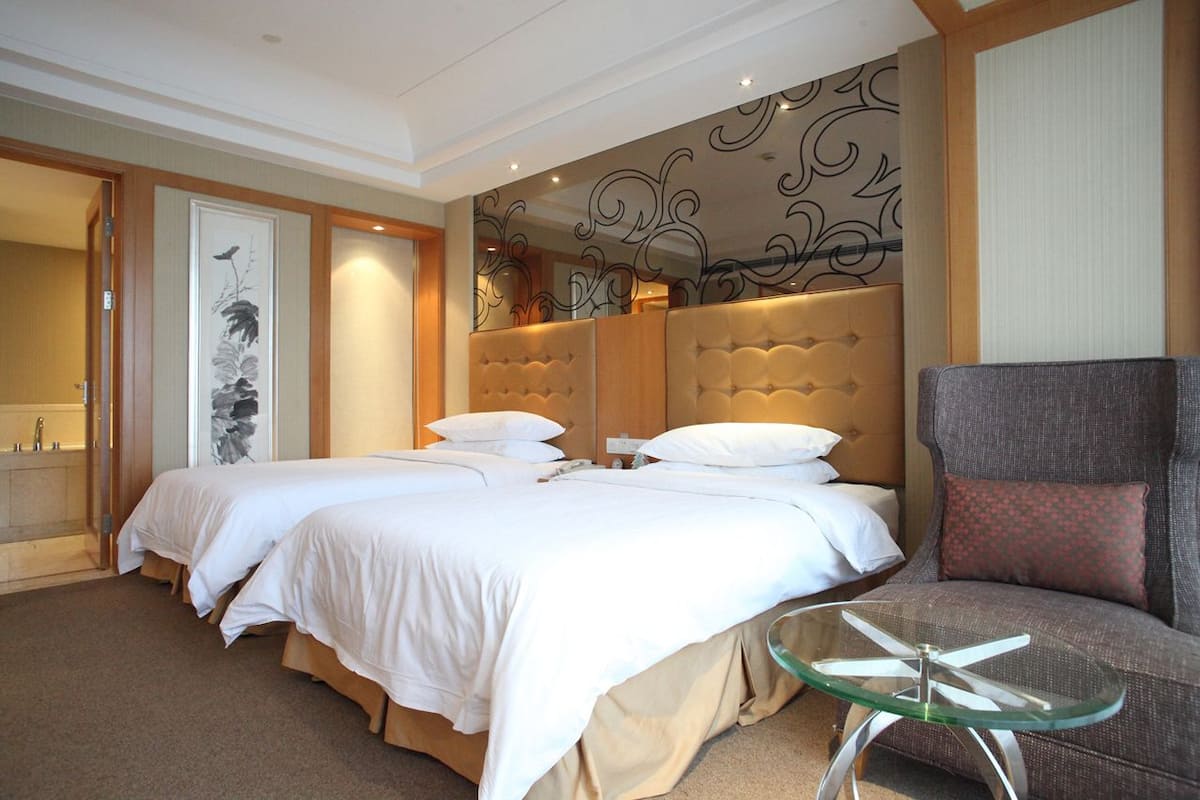 Une chambre d’hôtel avec deux lits, un coin salon comprenant un fauteuil, une table en verre et un panneau décoratif mural.