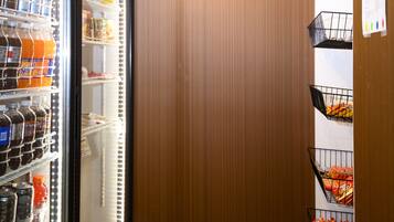 A corner of a room with a refrigerator stocked with beverages, a wooden panel wall, and a shelving unit with snacks.