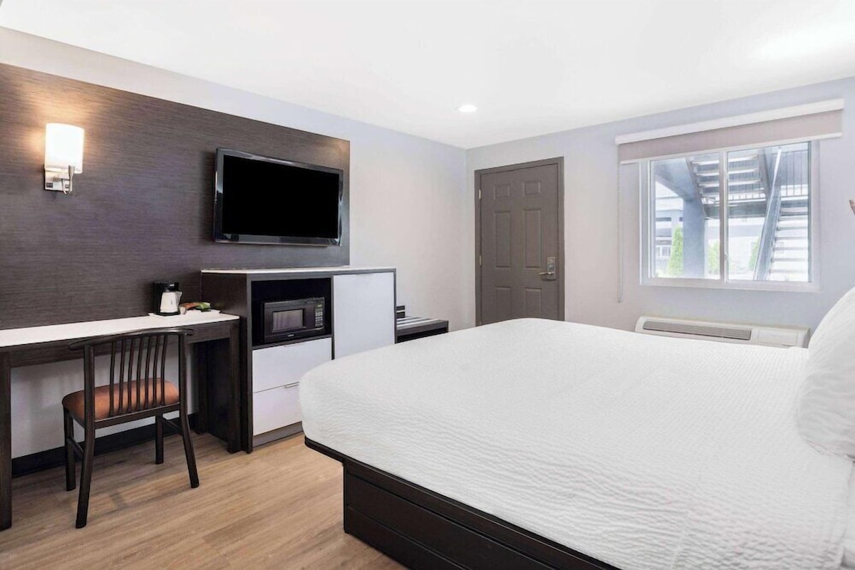 A hotel room with a bed, a desk with a chair, a flat-screen TV, a microwave, and a window with a view of a building.