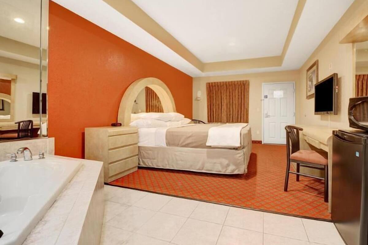 A hotel room with a bed, a desk, a chair, a TV, a mirror, a door, and a bathroom with a bathtub and sink.