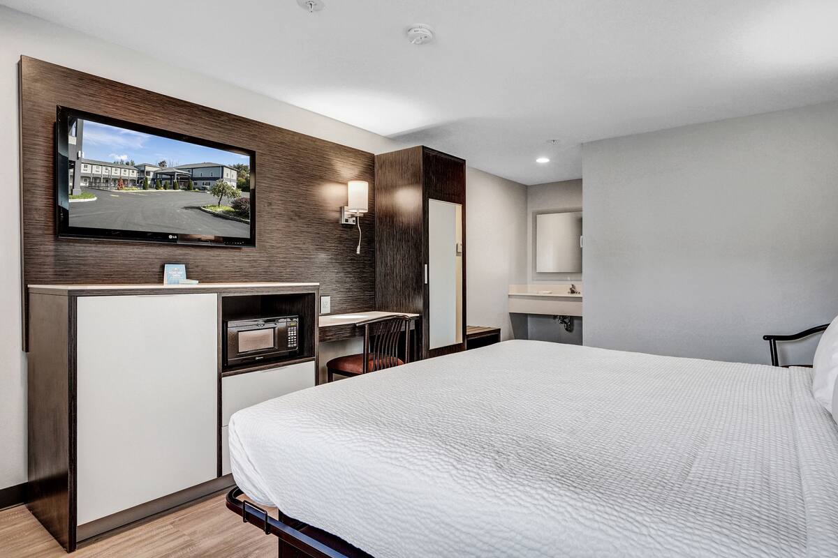 A hotel room with a bed, a desk with a microwave, a TV, and a bathroom with a mirror and sink.