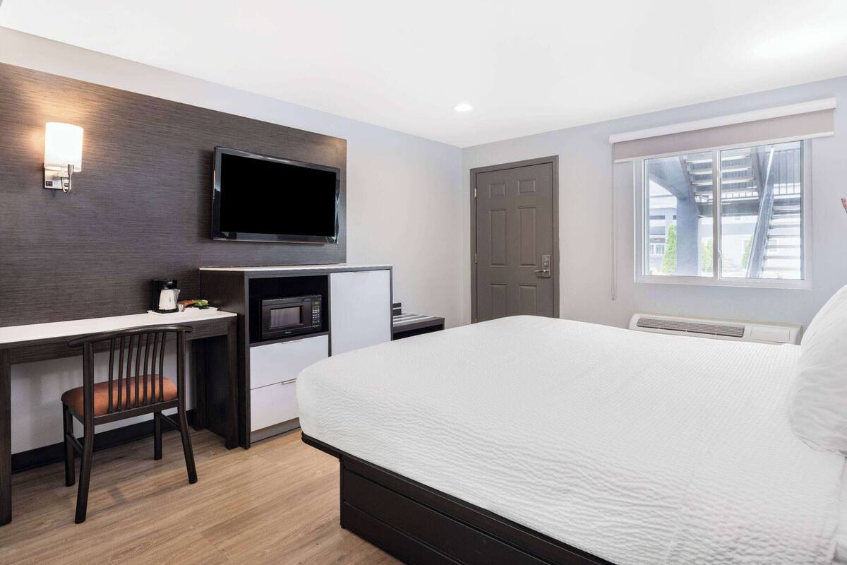 A hotel room with a bed, a desk with a chair, a flat-screen TV, a microwave, and a window with a view of buildings.