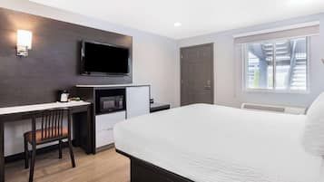A hotel room with a bed, a desk with a chair, a flat-screen TV, a microwave, and a window with a view of buildings.