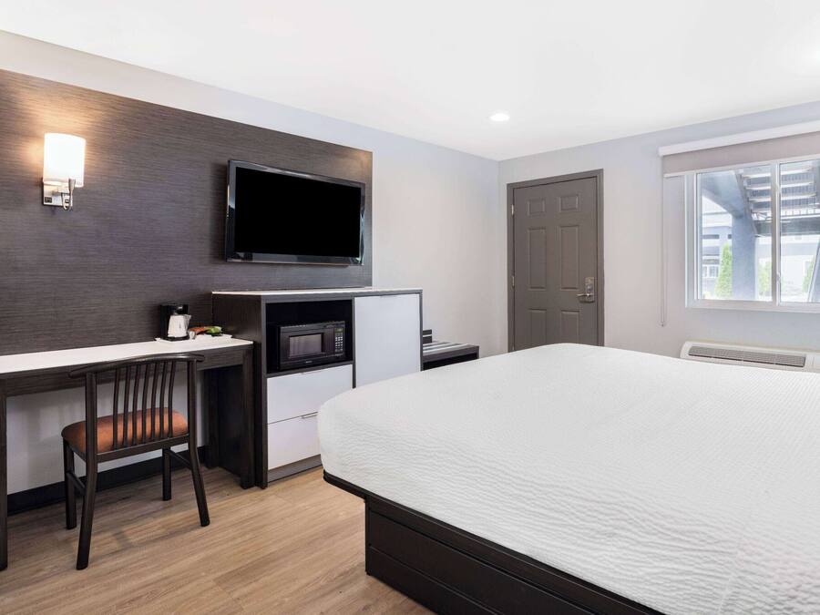 A hotel room with a bed, a desk with a chair, a flat-screen TV, a microwave, and a window with a view of buildings.