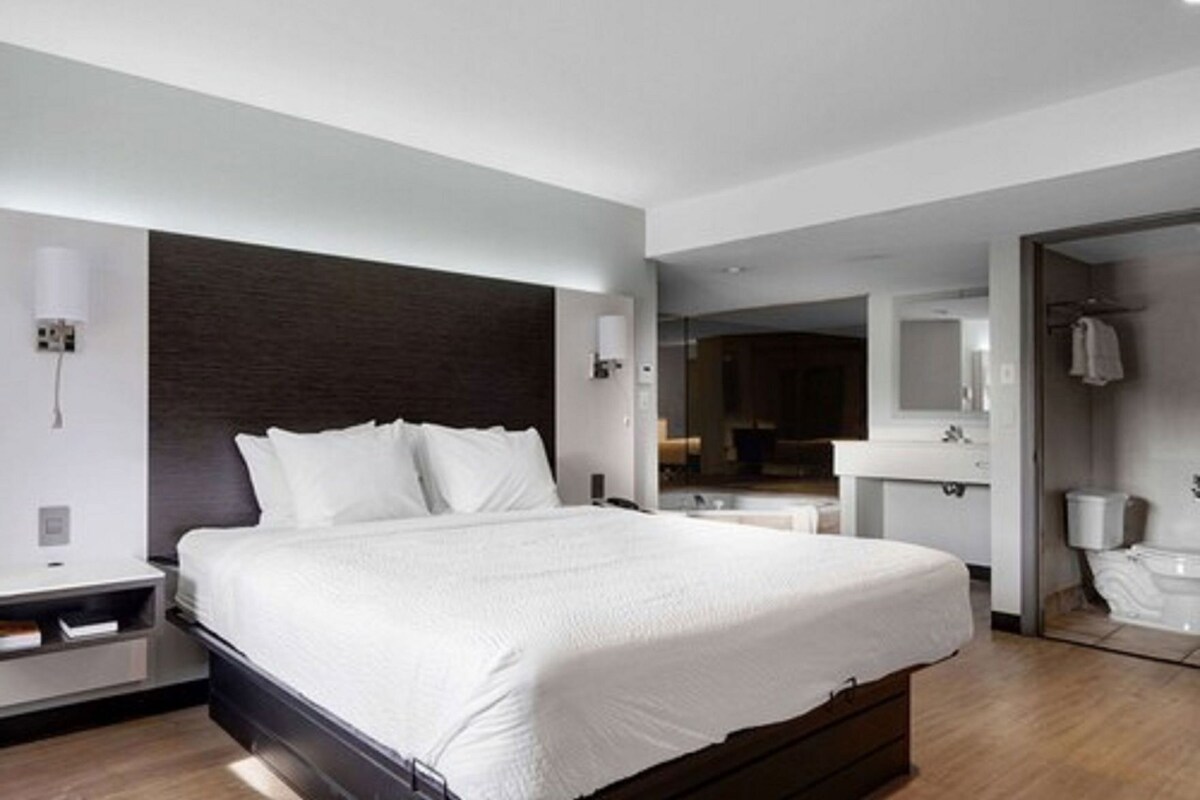 A modern hotel room with a large bed, a bedside table, a mirror, and a bathroom visible through an open door.