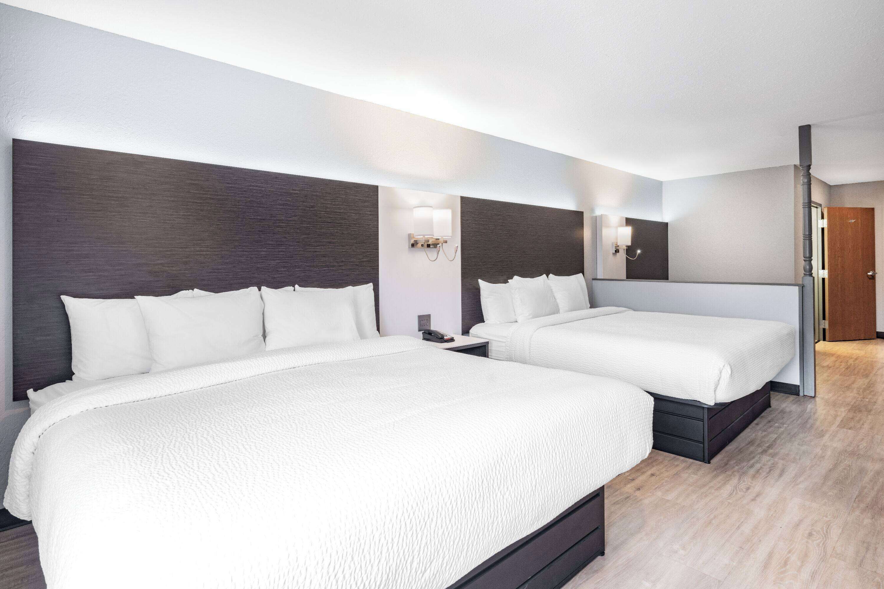 A hotel room with two beds, dark wood headboards, white bedding, a nightstand with a lamp, and a wooden door.