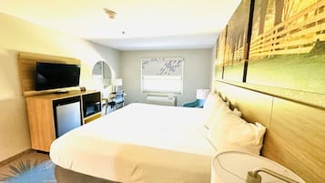 Habitación de hotel con cama, escritorio con computadora, televisión, ventana con persianas y una obra de arte colgada en la pared.