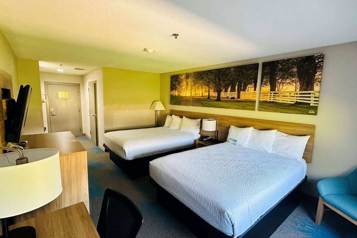 A hotel room with two beds, a desk with a lamp, a chair, and a large window with a painting of a park scene.