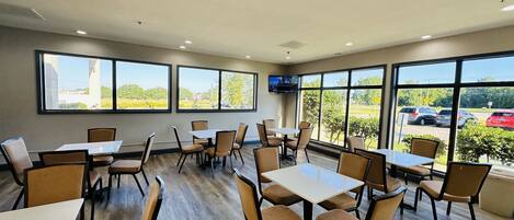 A spacious indoor dining area with large windows, multiple tables and chairs, and a flat-screen TV mounted on the wall.