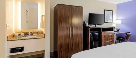 Una habitación de hotel con cama, armario de madera, televisor, escritorio con lámpara y baño con lavamanos y espejo.