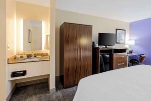 Una habitación de hotel con cama, armario de madera, televisor, escritorio con lámpara y baño con lavamanos y espejo.