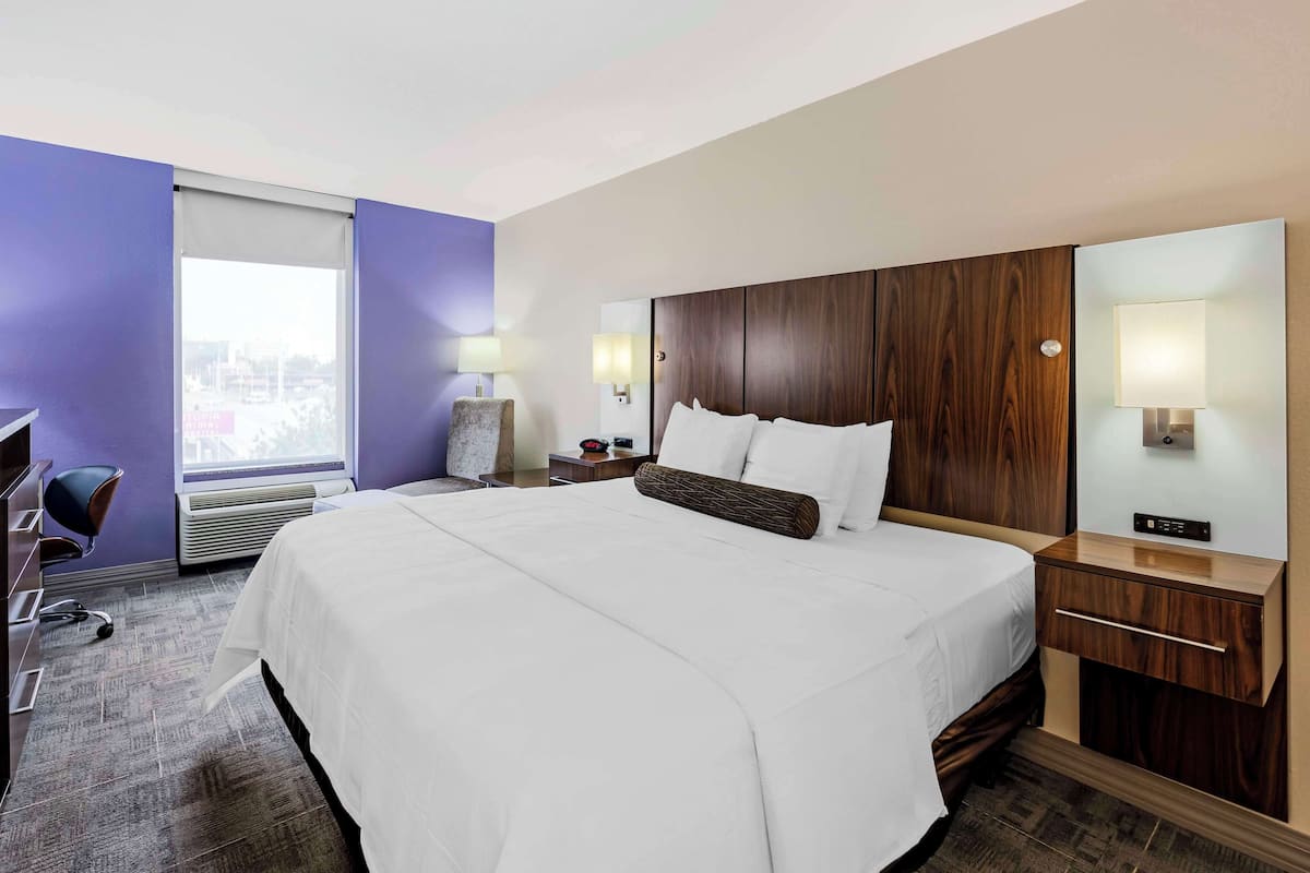 A hotel room with a large bed, a desk, a chair, a window with blinds, and a city view.