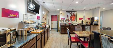 Cocina moderna con microondas, tostadora y exhibición de alimentos. Hay un comedor con sillas rojas y una mesa.