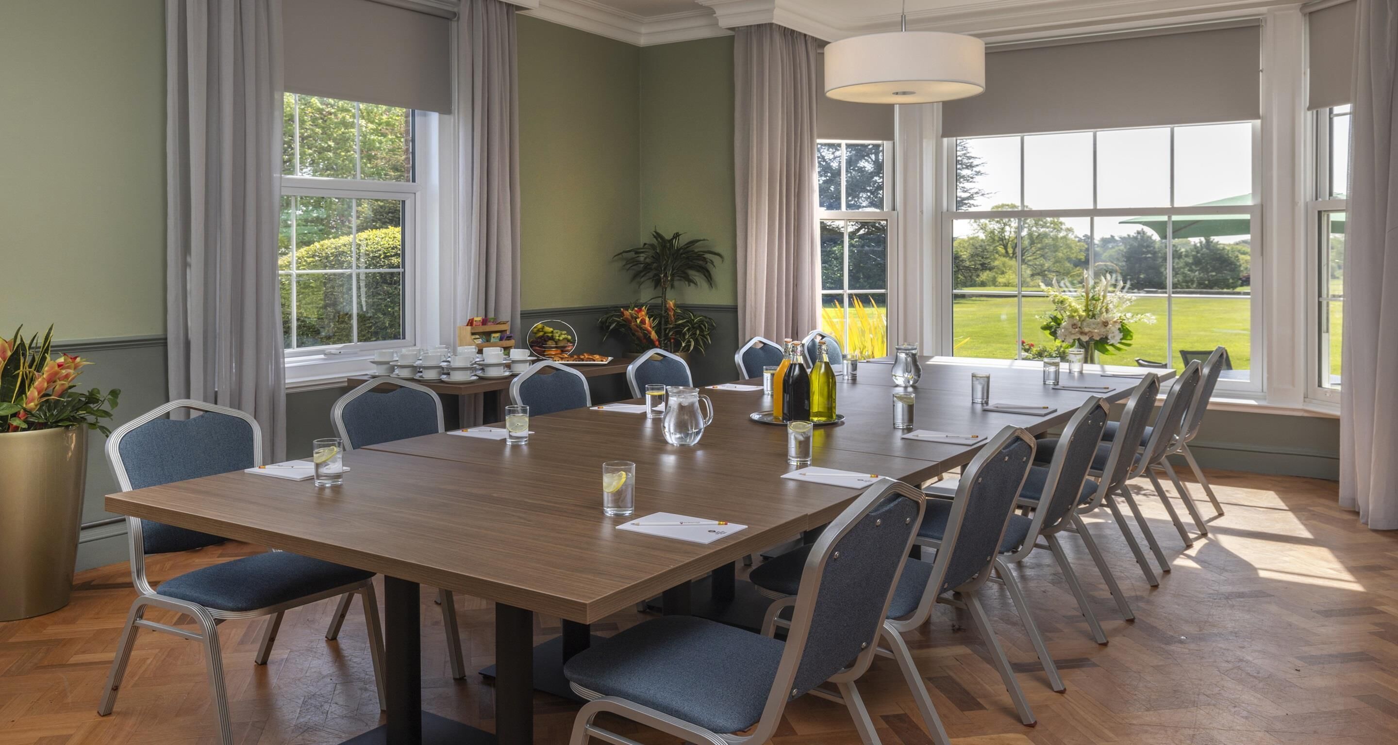 A conference room with a long table, chairs, a potted plant, and a view of a green landscape through large windows.