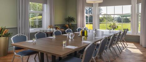 A conference room with a long table, chairs, a potted plant, and a view of a green landscape through large windows.