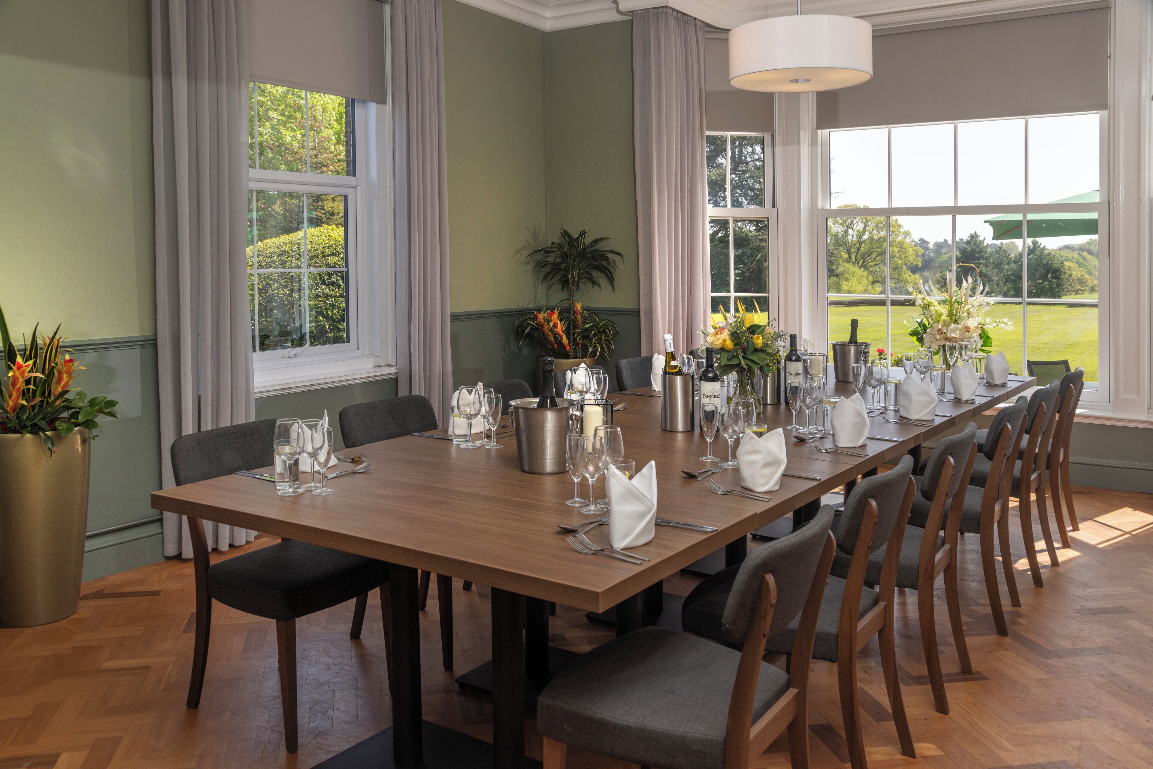 A dining room with a long wooden table set for a meal, large windows with curtains, and a view of the outdoors.