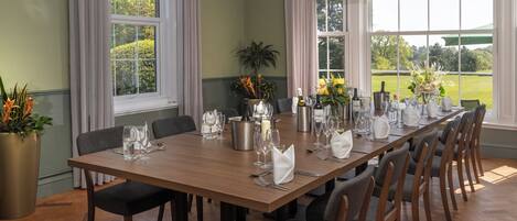A dining room with a long wooden table set for a meal, large windows with curtains, and a view of the outdoors.