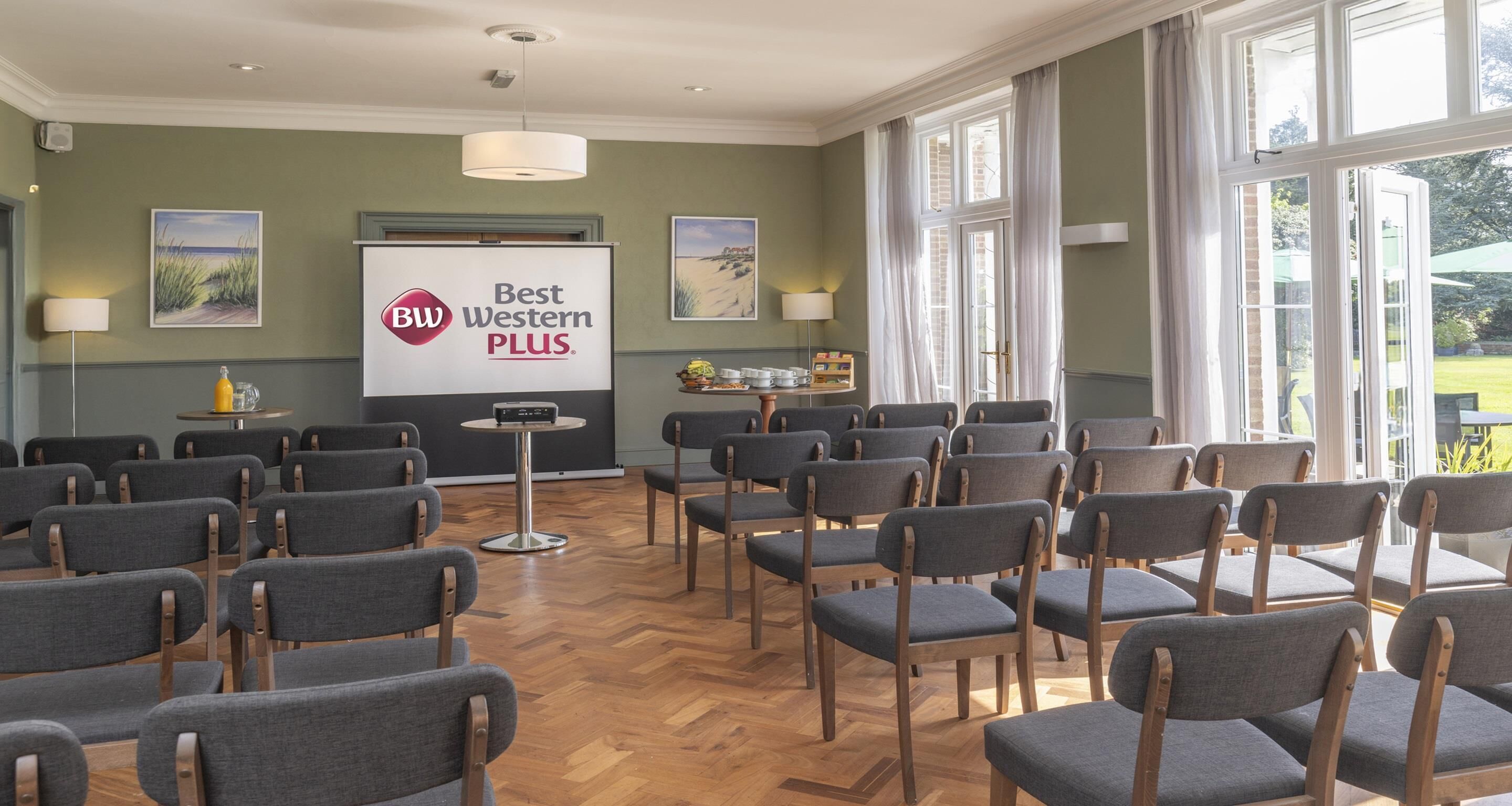 A conference room with rows of chairs, a projector screen displaying "Best Western PLUS", and a large window with curtains.