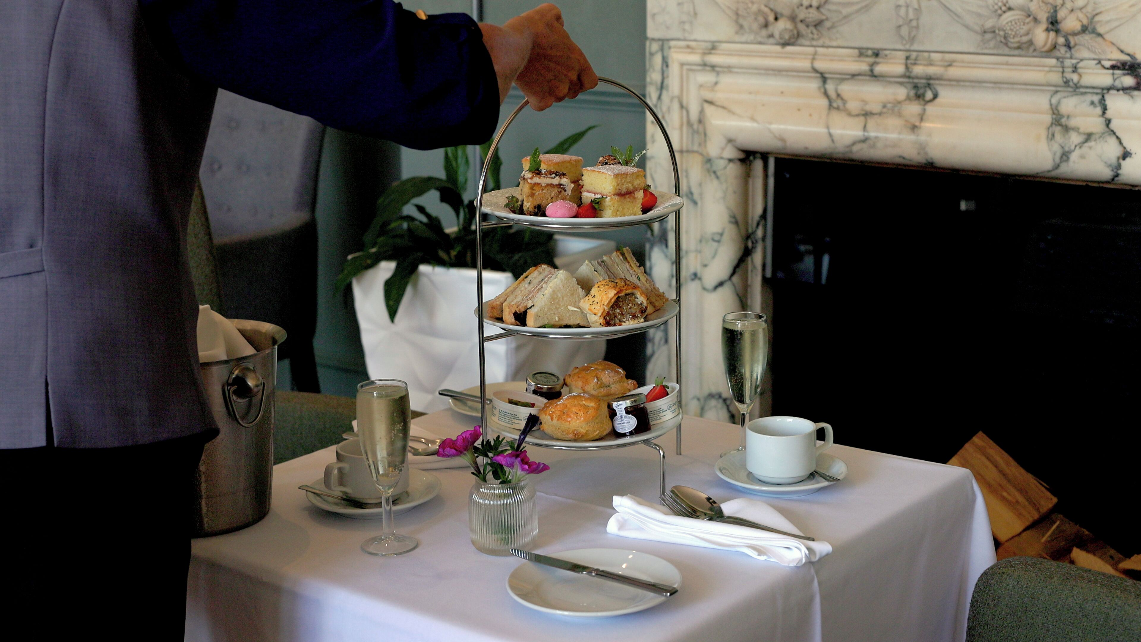 A tiered tray of sandwiches and pastries on a table with a white tablecloth, a glass of water, and a cup of tea.