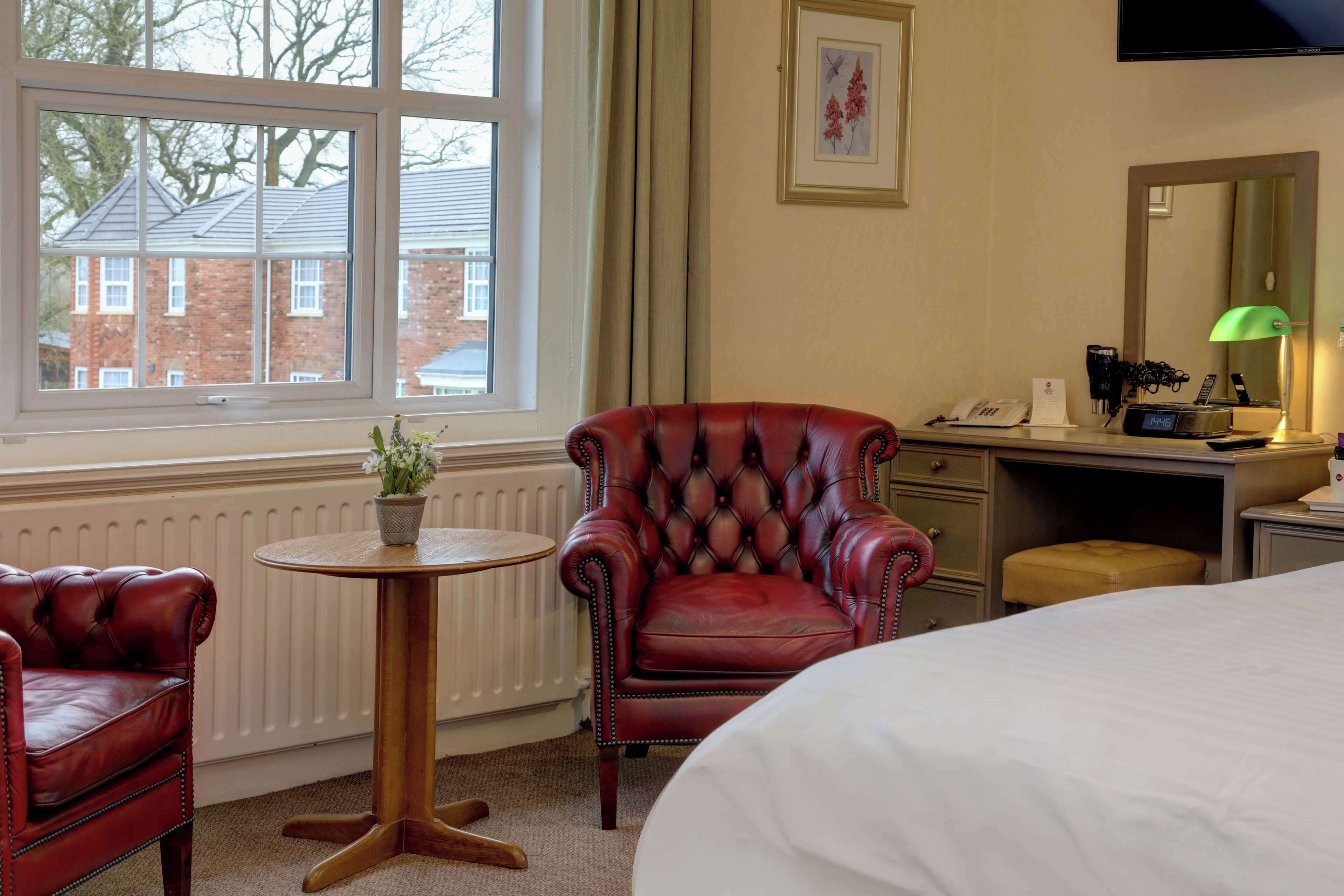 Ein Hotelzimmer mit einem Bett, einem roten Ledersessel, einem kleinen runden Tisch mit Blumentopf, einem Schreibtisch mit Lampe und einem Fenster mit Blick auf Gebäude.