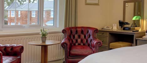 A hotel room with a bed, a red leather armchair, a small round table with a potted plant, a desk with a lamp, and a window with a view of buildings.