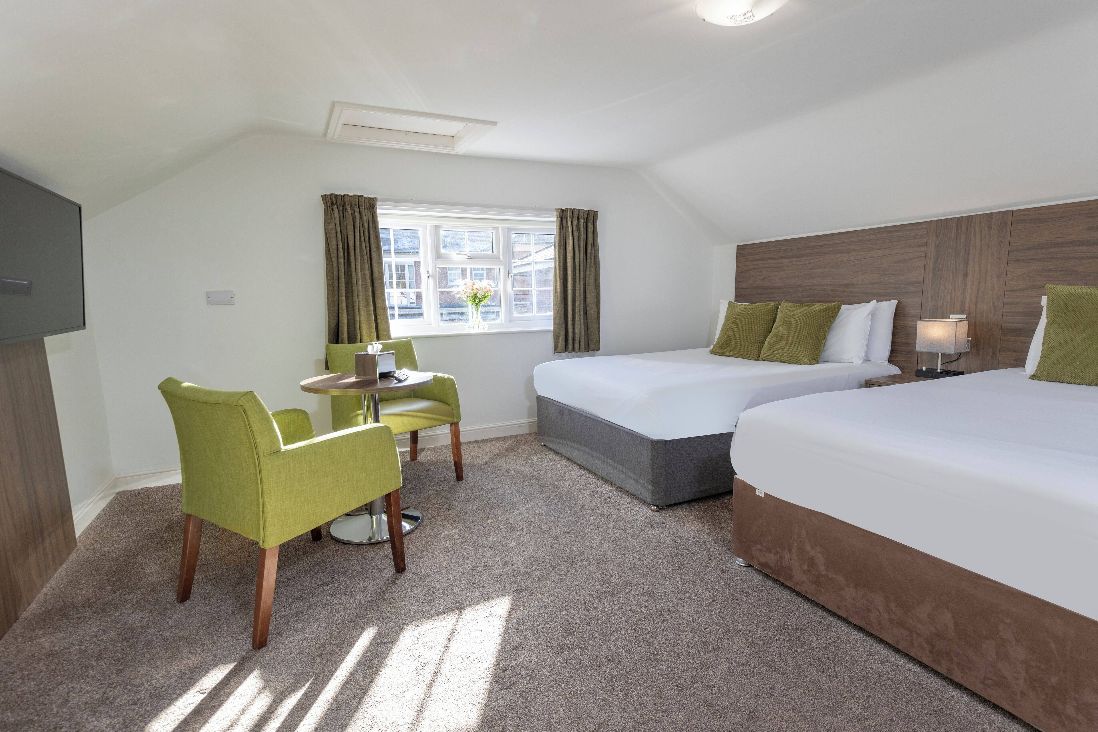 Habitación de hotel con dos camas, zona de comedor con mesa y sillas, ventana con cortinas y televisión de pantalla plana.