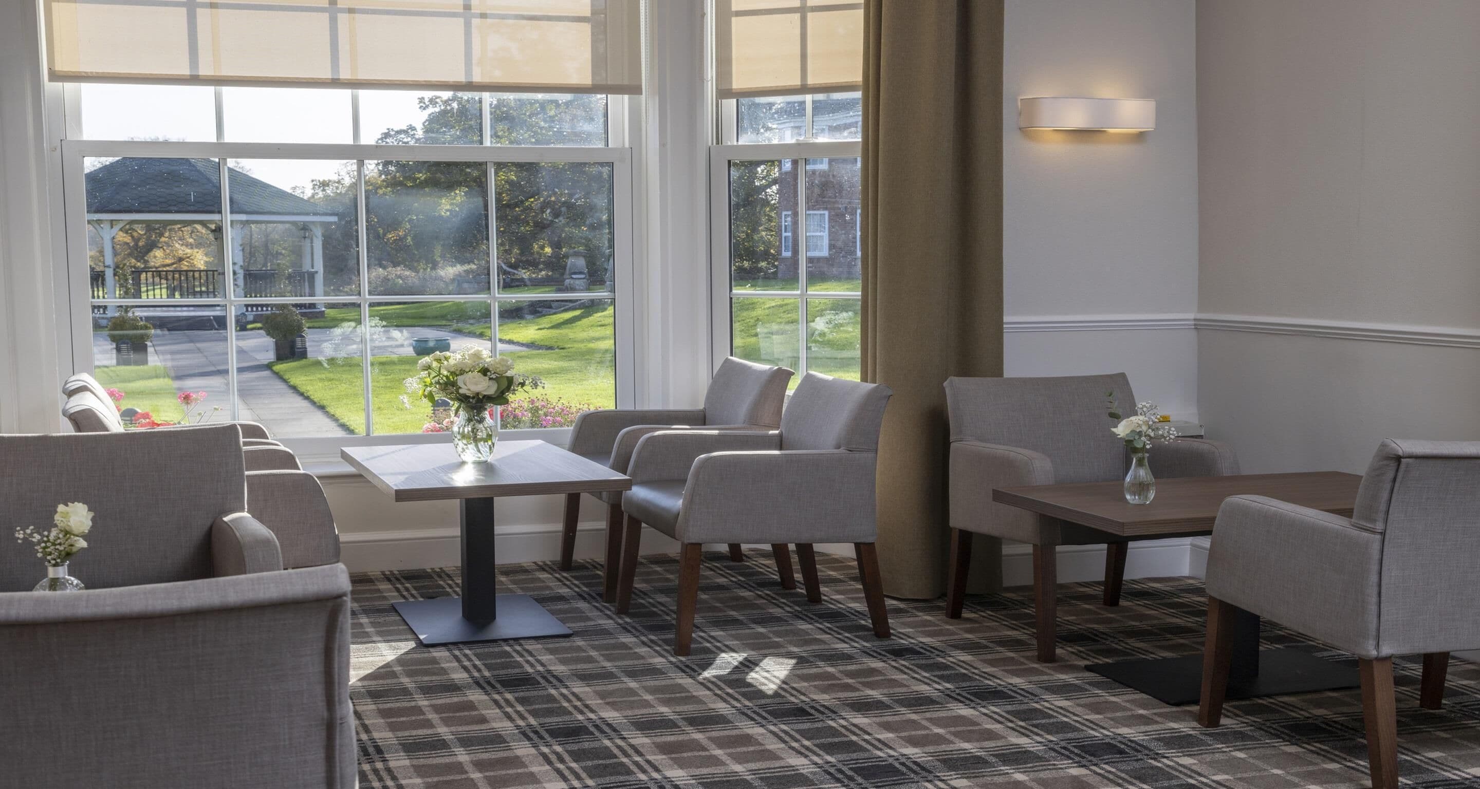 A room with a dining table, four chairs, a small side table, and a view of a garden through large windows.